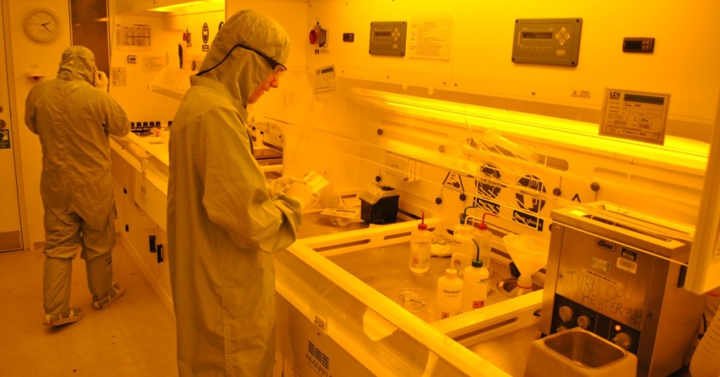 Scientists working in an orange-lit photolithography cleanroom designed to protect light-sensitive photoresist.