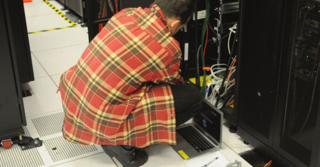 Technician working with a laptop beside physical server racks, representing the physical hardware that hosts virtual machines.