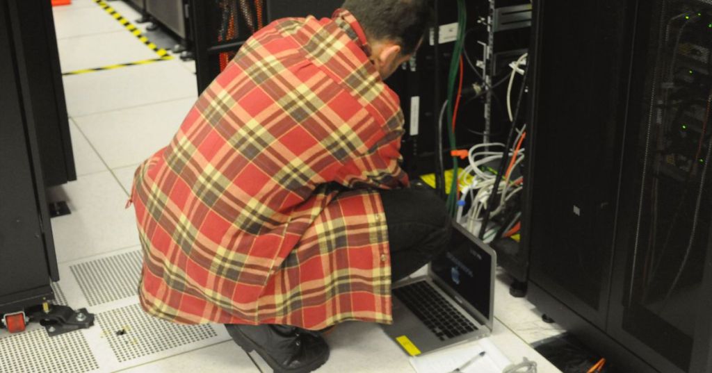 System administrator working with a laptop beside server racks in a data center.