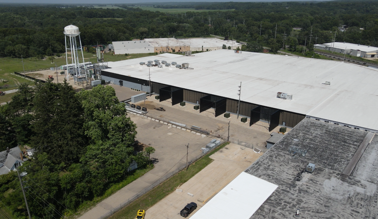 July 2026 aerial view of the real Dowagiac Hyperscale Data facility, with broad factory roofs, utility equipment and a water tower next to wooded neighborhood