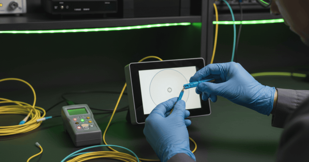 Fiber optics technician inspecting and cleaning an LC connector beside an optical power meter and light source on a professional test bench.