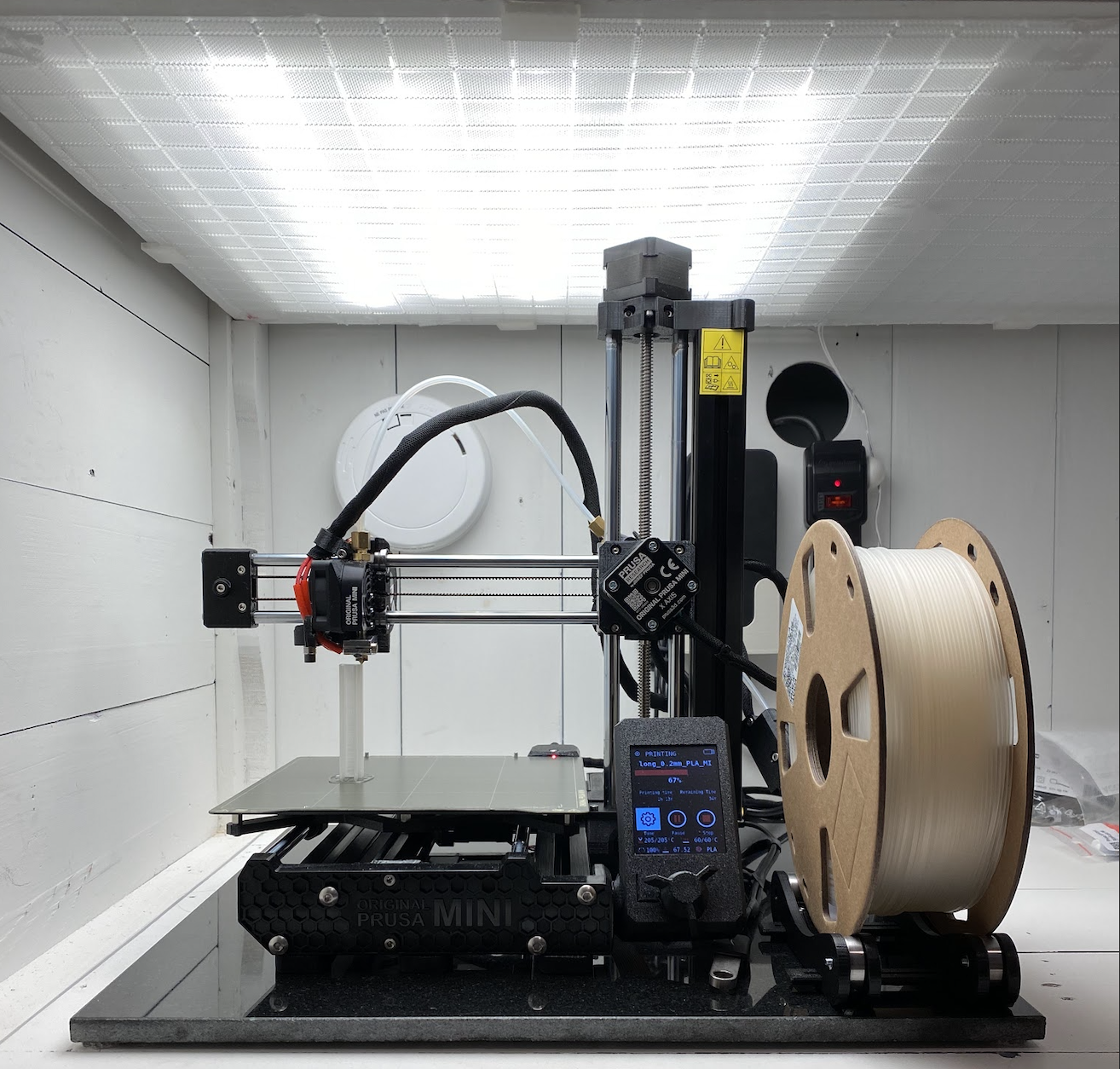 A Prusa Mini 3D printer with a spool of white filament in a compact makerspace.