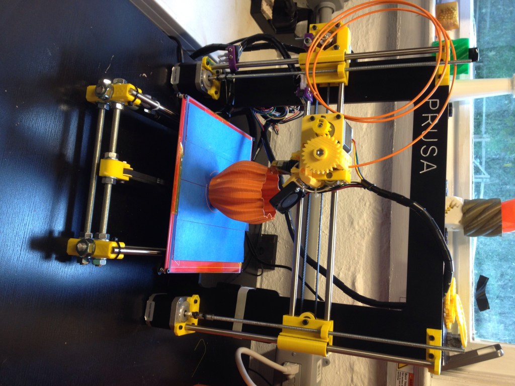A Prusa i3 RepRap 3D printer building an orange vase layer by layer.