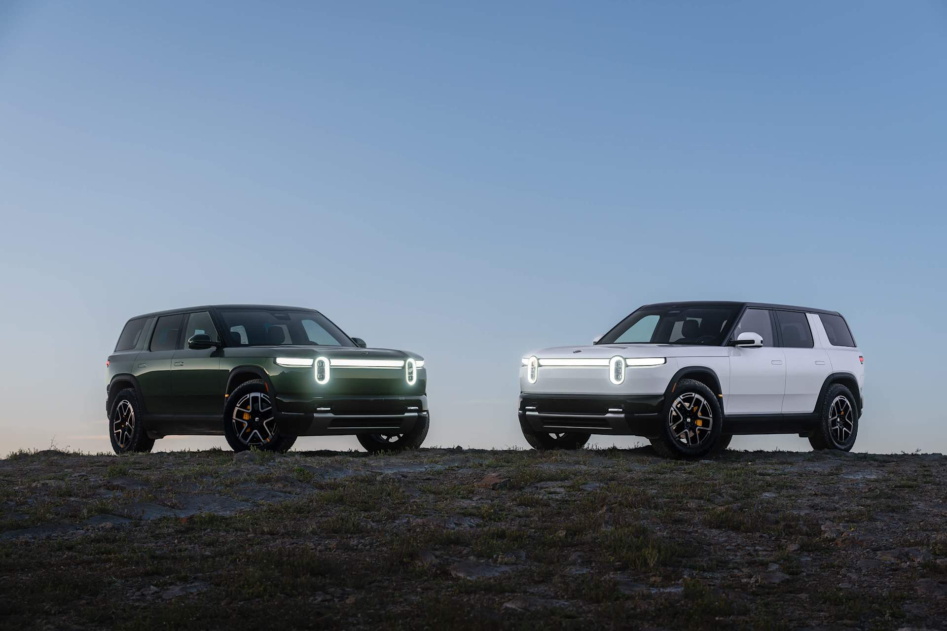 Two Rivian R2 electric SUVs in green and white on a hillside at dusk