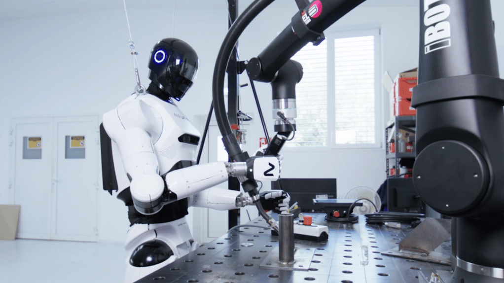 A NEURA humanoid robot working beside a robotic arm in a robotics laboratory.